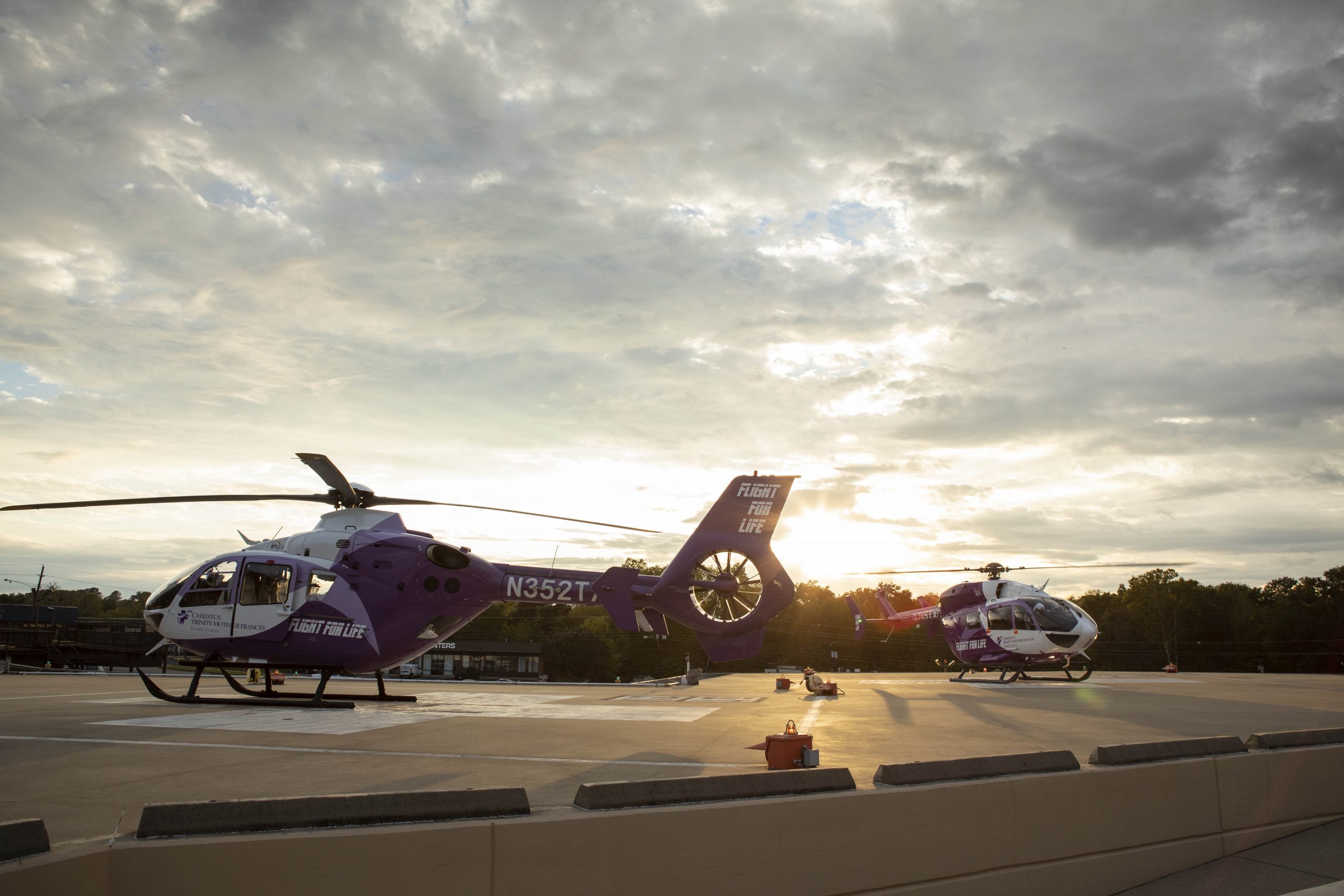 Two medical helicopters sit on a landing pad as the sun sets in the background.