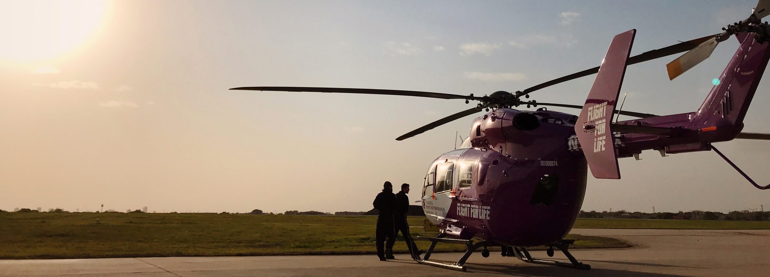 A Flight For Life helicopter sits parked on a runway tarmac, with two crew members next to it.