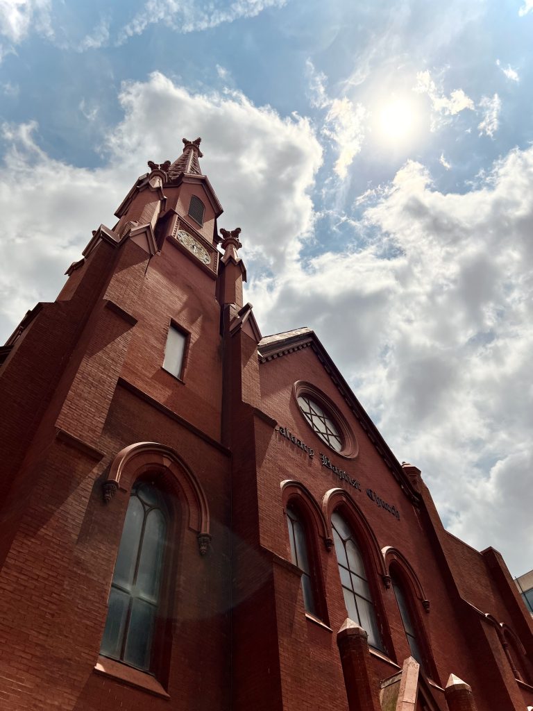 A red brick church, viewed from a low angle, with a cloudy sky behind.