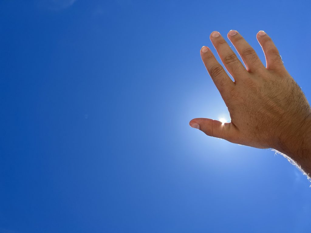 A hand reaches up to the bright, blue sky, blocking the bright sun.
