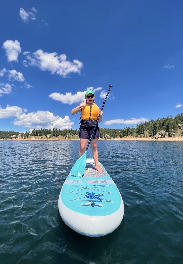 A woman stands on a stand up paddle board on a blue lake.