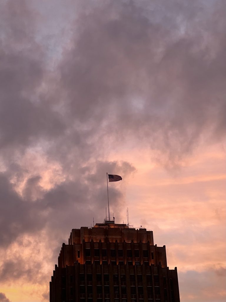 A tall building with a waving American flag in front of a pink and orange sunset clouds.