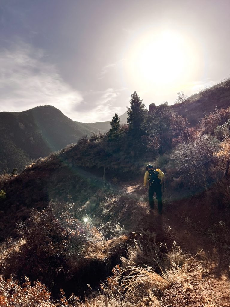 A wildland firefighter with a load pack and hand tool walks up a trail in dry, mountainous terrain.