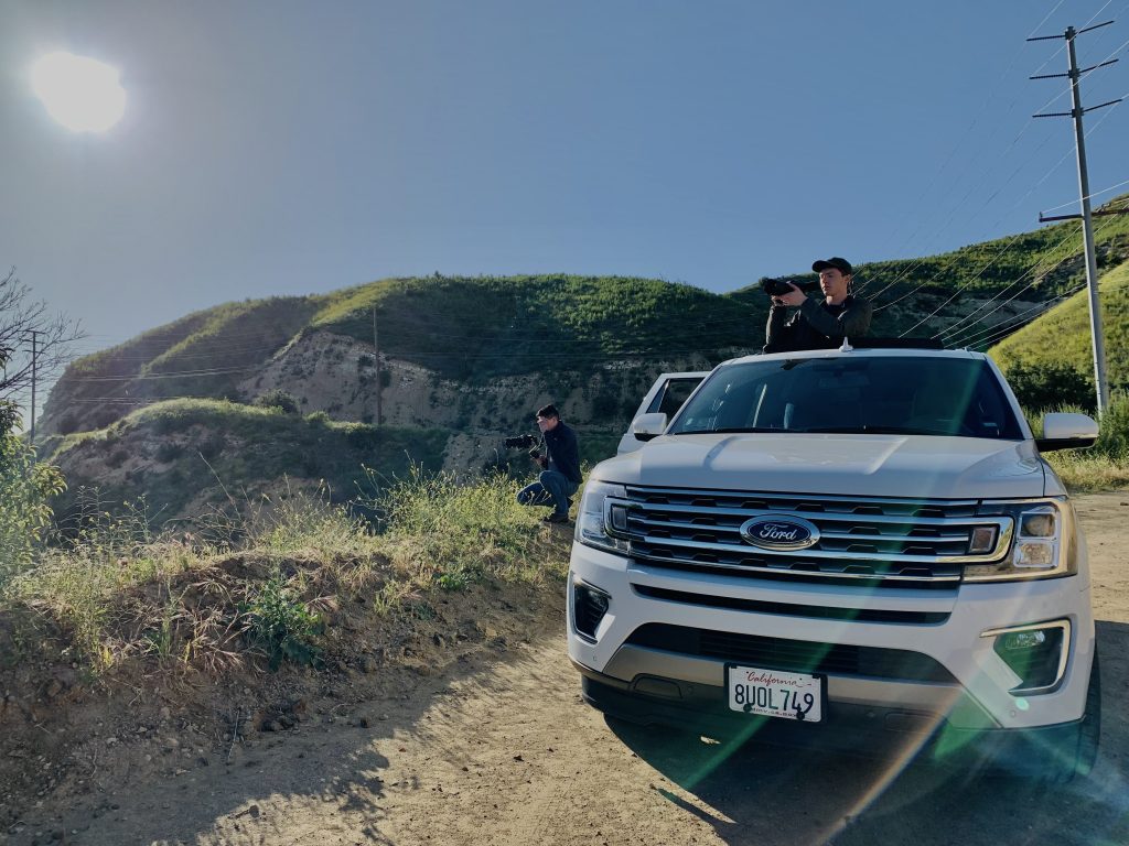 A Ford SUV is parked in gravel along the side of a California highway, with two individuals shooting video and photos.