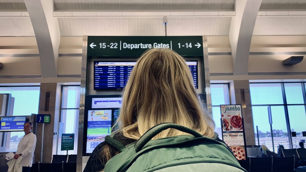 A blonde-haired female looks at an airport departures board, with arrows pointing to gates to the left and right.