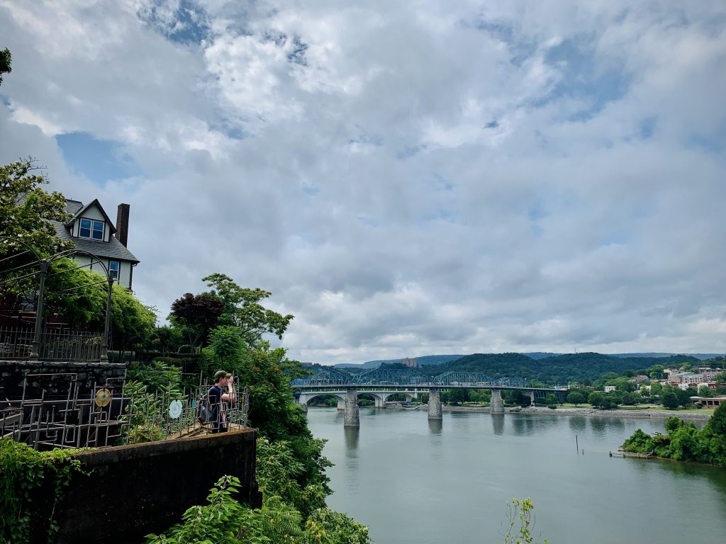 A wide open view of Chattanooga, Tennessee, bridges crossing the Tennessee River, and lush rolling hills behind. In the foreground, a photographer leans over a balcony to take a picture.