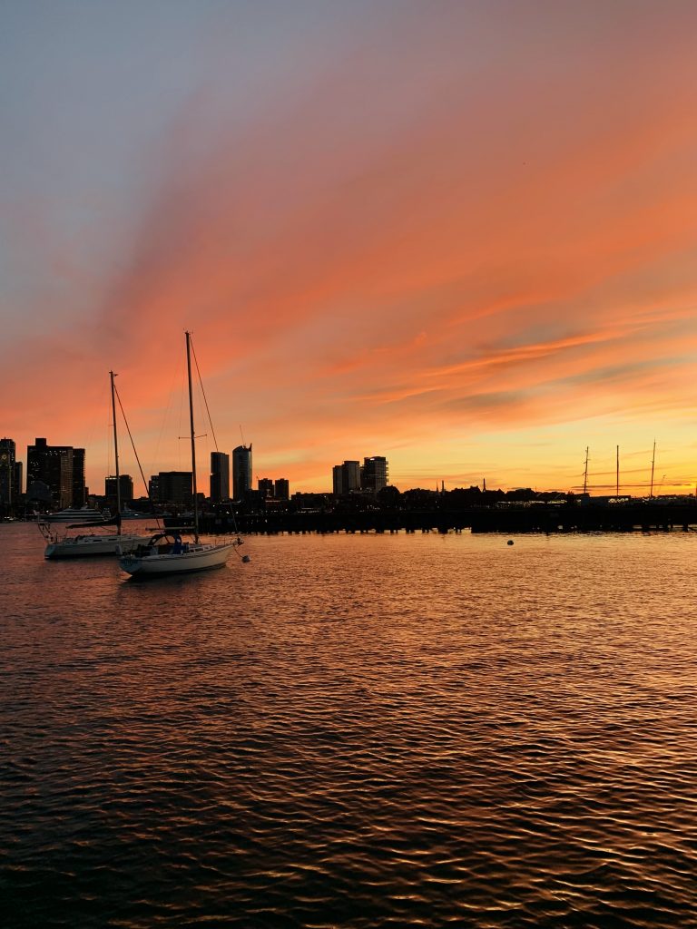 Two sailboats bob in the foreground as fiery sunset clouds create a silhouette of the Boston skyline.
